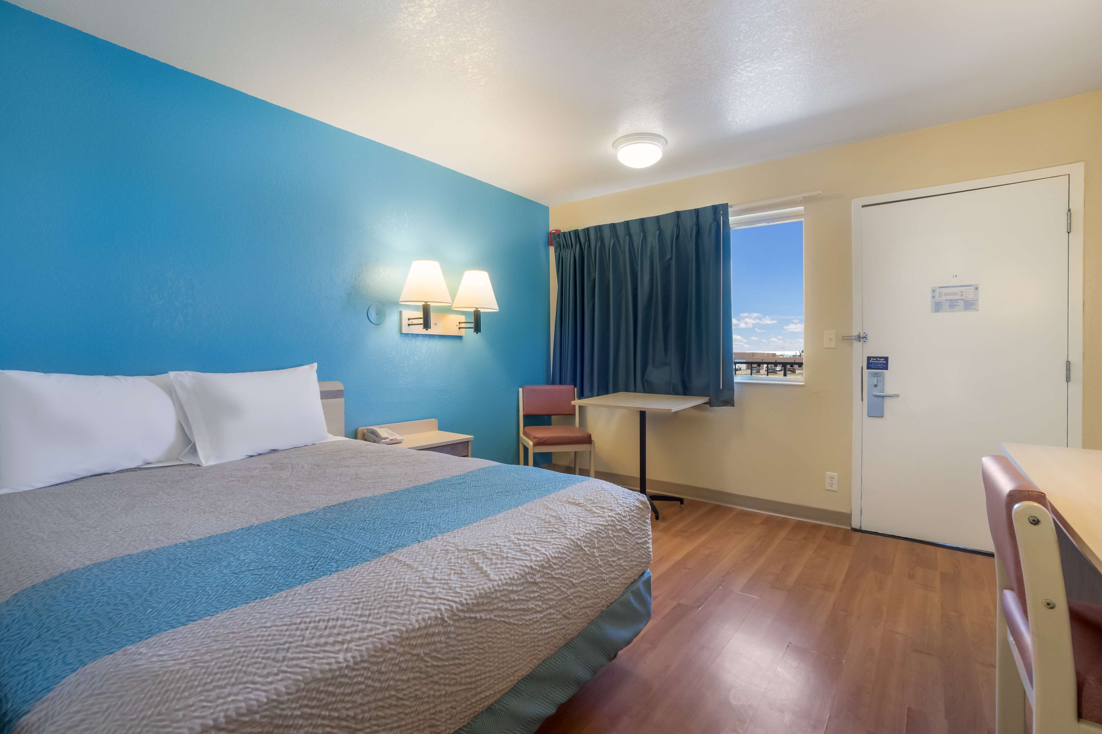 A hotel room with a bed, a desk, a chair, a window with curtains, and a door.