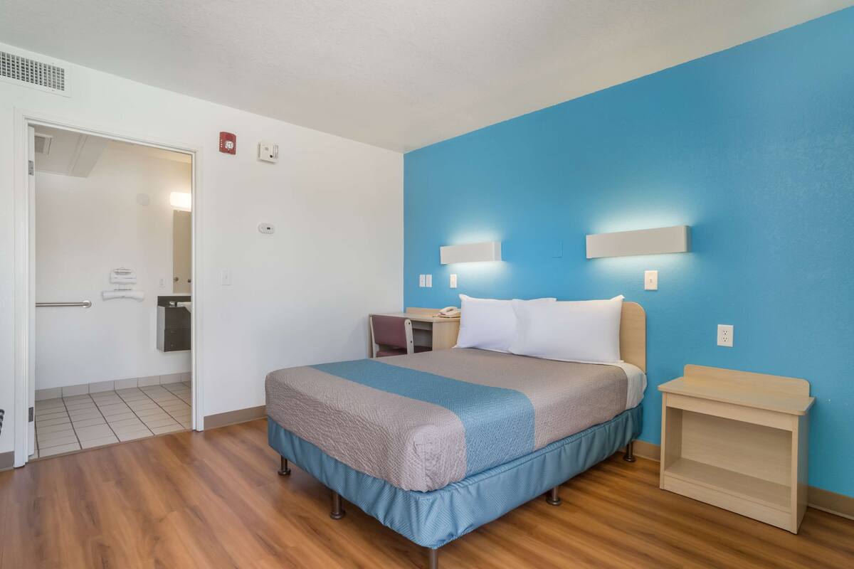 A hotel room with a bed, wooden flooring, a blue accent wall, a nightstand, and a door leading to a bathroom.