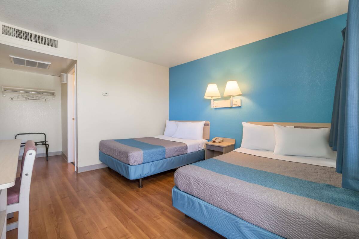 A hotel room with two beds, a desk, and a chair. There are two wall-mounted lamps and a ceiling vent.