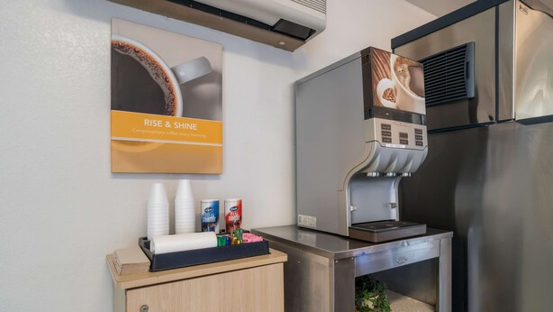 A modern kitchen with a coffee machine, a stainless steel refrigerator, and a wooden cabinet with cleaning supplies. - Motel 6 Grand Junction, CO (Grand Junction)