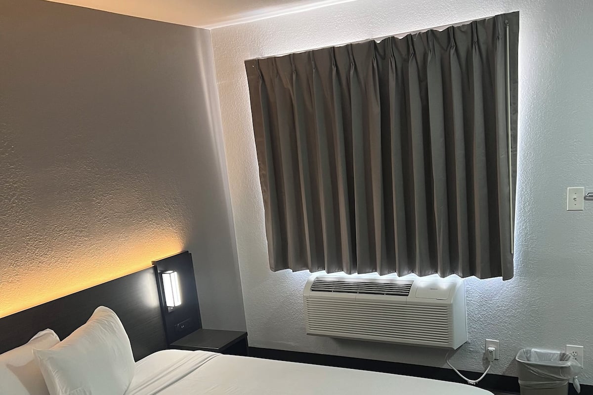 A hotel room with a large bed, a nightstand, a wall-mounted air conditioner, and a window with curtains.
