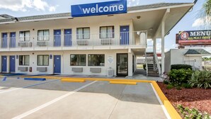 Un motel con un letrero de 'Welcome', un área de estacionamiento techada y un cartel de 'Menu' que anuncia comidas a menos de $4.