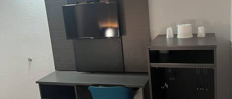 Una habitación moderna con un escritorio, una silla azul, un televisor montado en la pared y un mueble bajo con tazas.