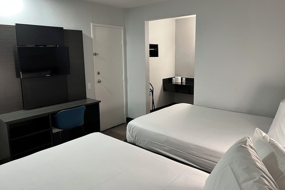 A hotel room with two beds, a desk with a chair, a TV, and a bathroom with a sink and mirror.