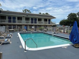Zona de piscina con sillones de descanso y una sombrilla, rodeada por una terraza cercada y un edificio con varias entradas.