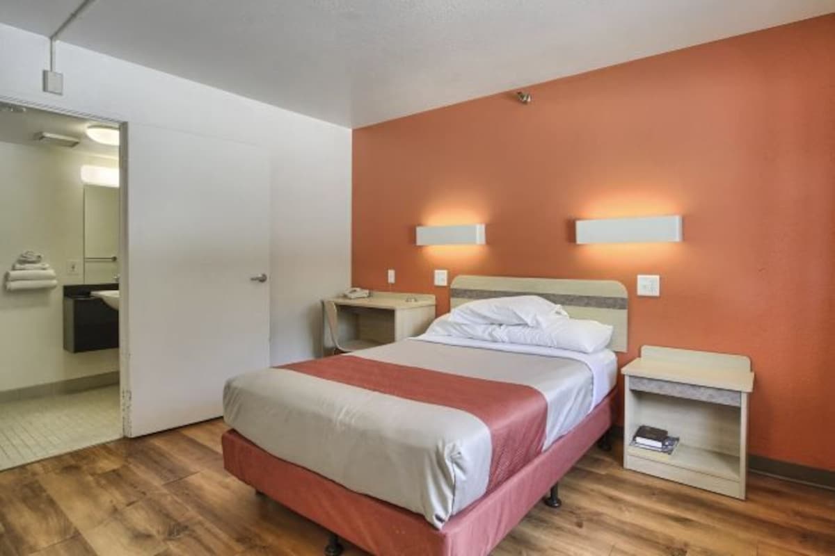 Une chambre d’hôtel avec un lit, des tables de chevet, une commode, un miroir et une salle de bain visible à travers une porte ouverte.