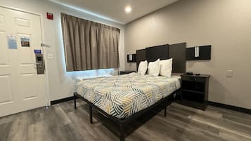 A modern hotel room with a bed, a nightstand, a window with curtains, a door, and a wall-mounted cabinet.
