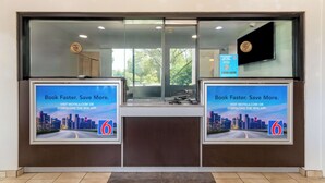 A reception desk with two large digital screens displaying a cityscape and promotional text for booking through a mobile app. - Motel 6 Rolling Meadows, IL - Chicago Northwest (Rolling Meadows)