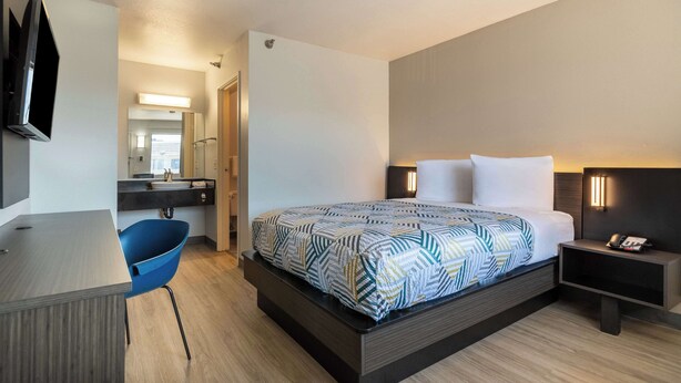 A modern hotel room with a bed, bedside table, chair, desk, and a TV mounted on the wall. - Motel 6 Rolling Meadows, IL - Chicago Northwest (Rolling Meadows)