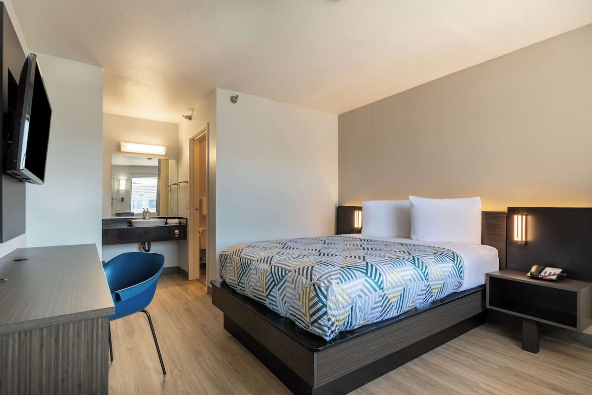 A modern hotel room with a bed, bedside table, chair, desk, and a TV mounted on the wall.