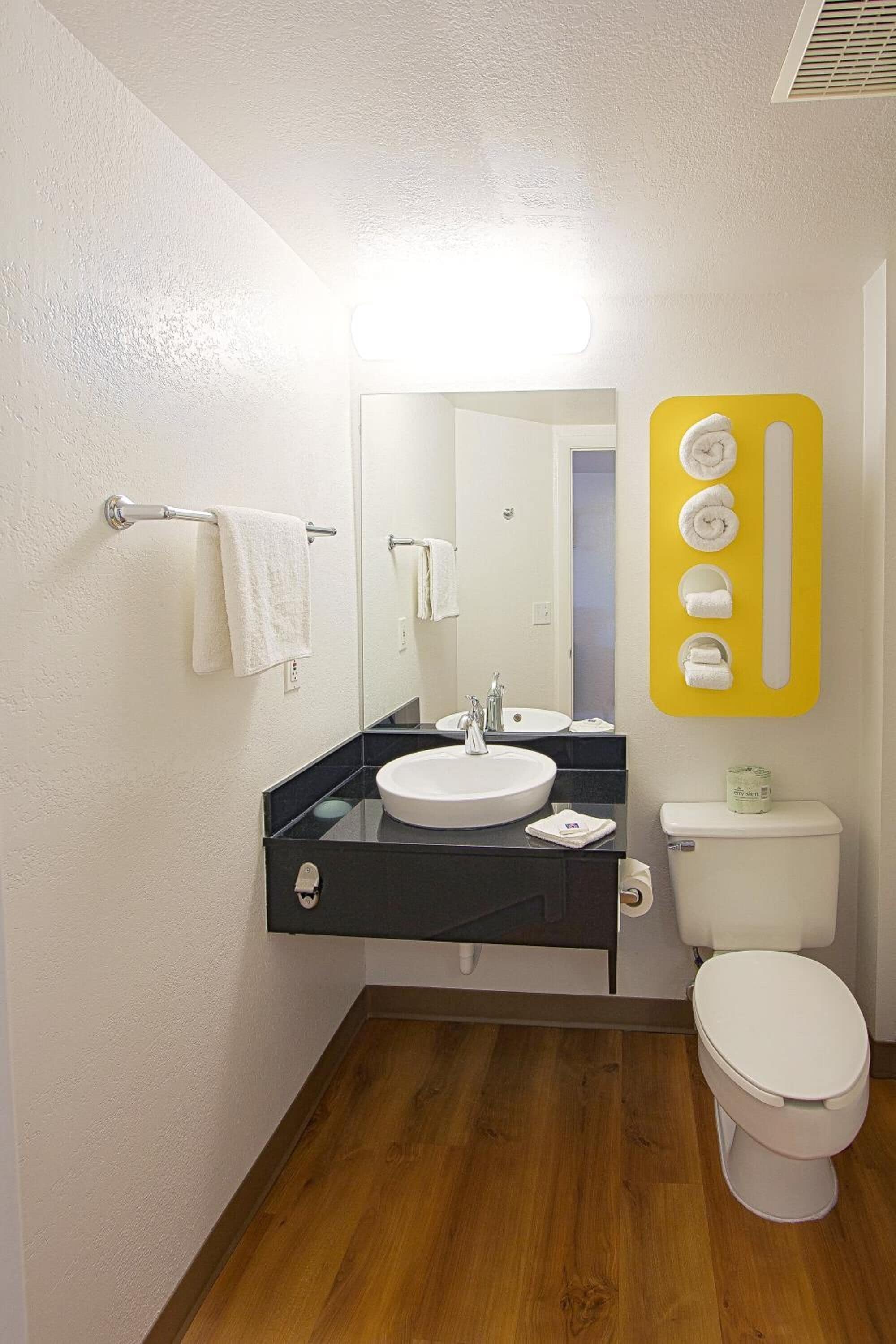 A bathroom with a white sink, mirror, and toilet. There is a towel rack with a white towel and a yellow towel holder with multiple towels.