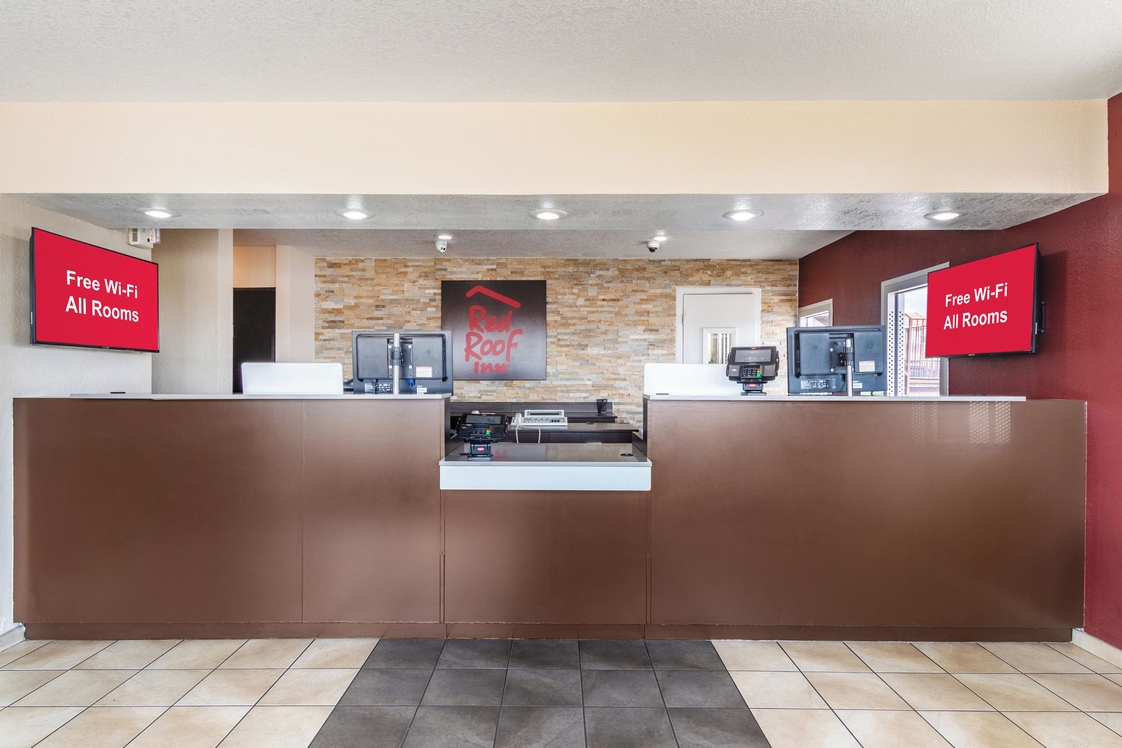 A hotel reception area with a stone wall backdrop, a Red Roof Inn sign, and two monitors displaying 'Free Wi-Fi All Rooms'.