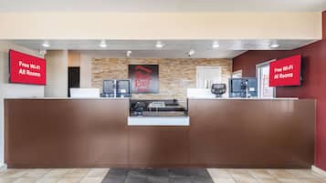 A hotel reception area with a stone wall backdrop, a Red Roof Inn sign, and two monitors displaying 'Free Wi-Fi All Rooms'.