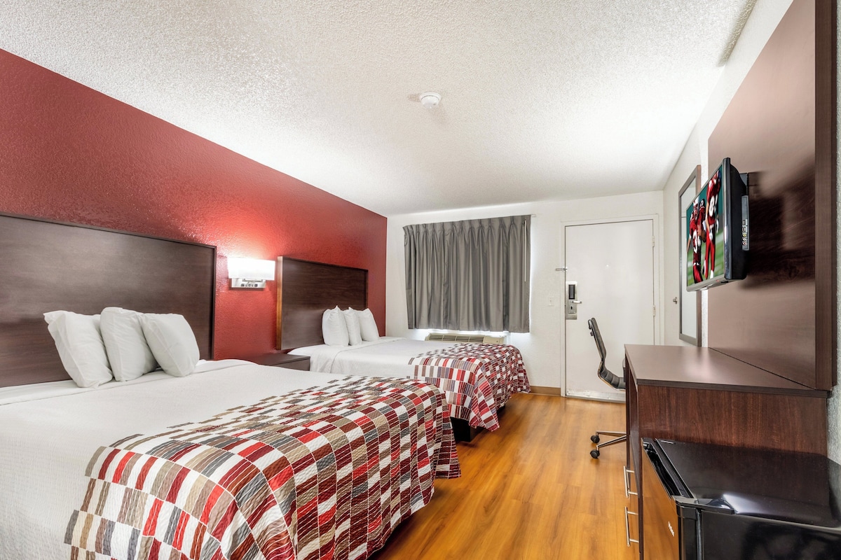 A hotel room with two beds, a desk, a TV, and a window with curtains.