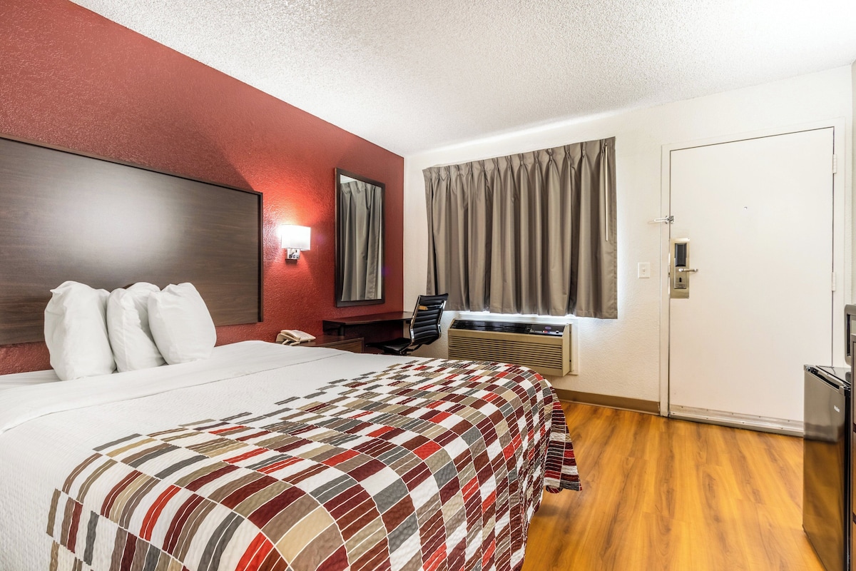 A hotel room with a bed, a desk, a chair, a window with curtains, a door, and a built-in air conditioning unit.