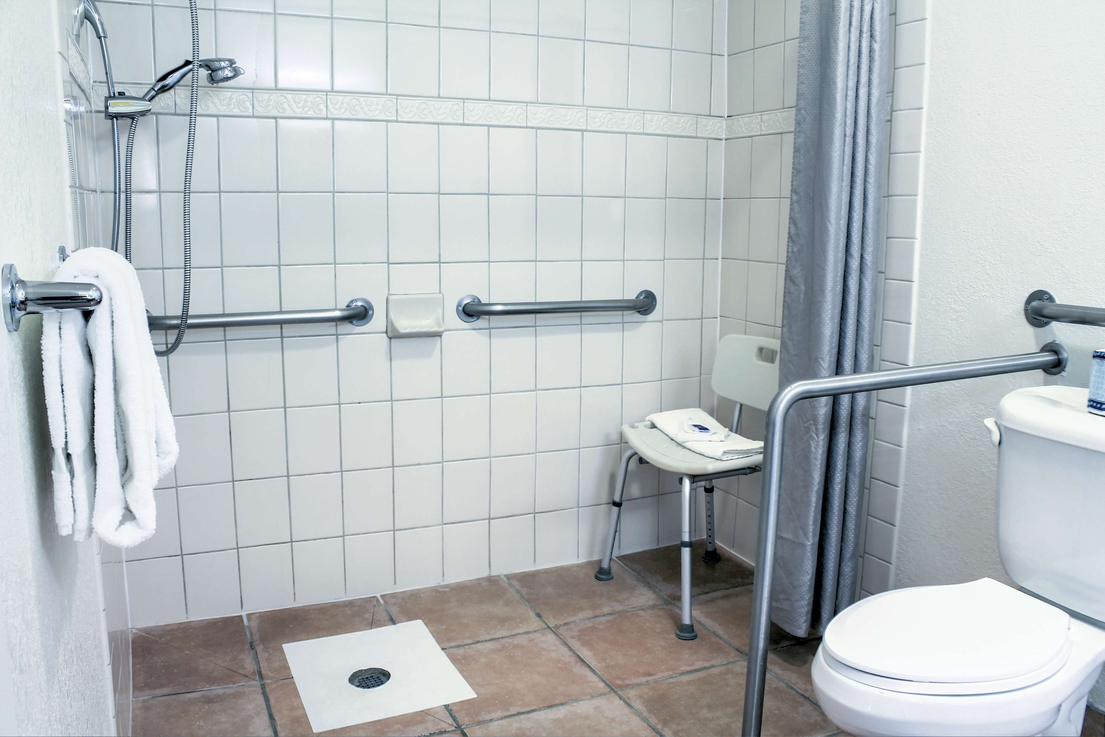 Et badeværelse med hvide flisemure, et hvidt toilet, en bruser med håndbruser og gelænder.