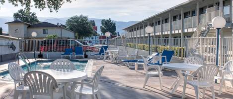 A pool area with white plastic chairs and tables, a pool, and a building with balconies in the background.