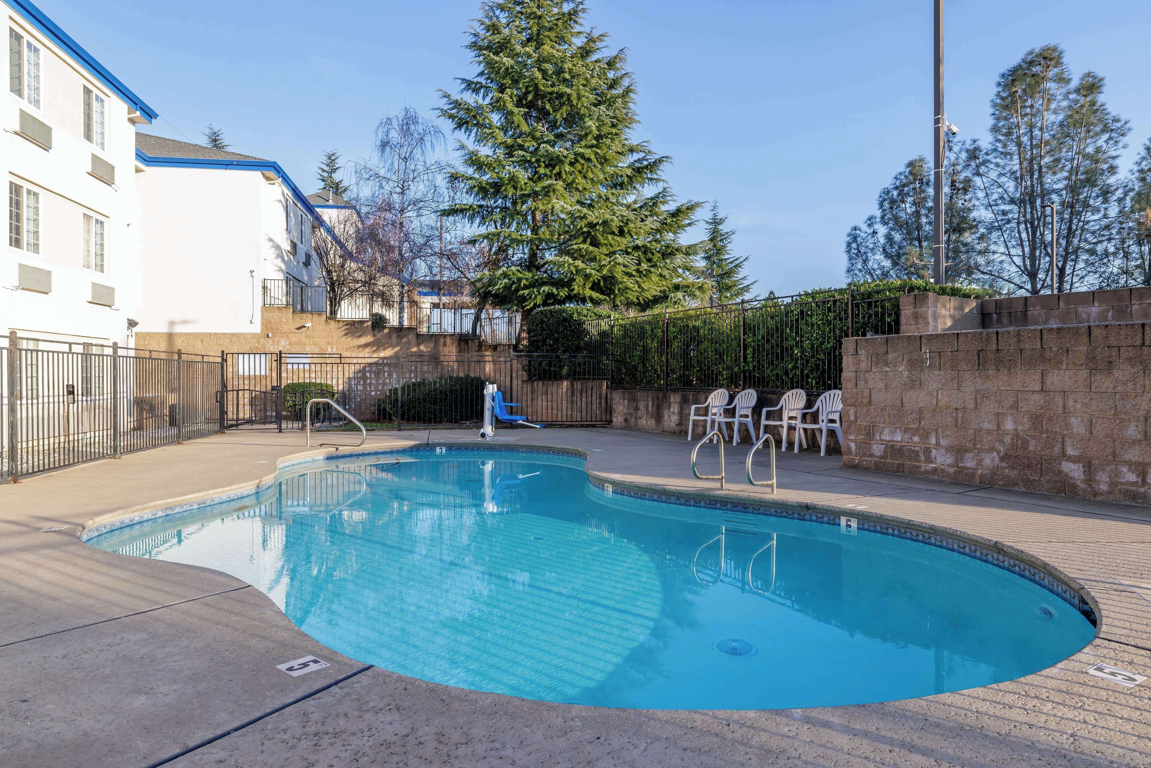 A swimming pool with a clear blue water, surrounded by a paved deck, a brick wall, and a metal fence.