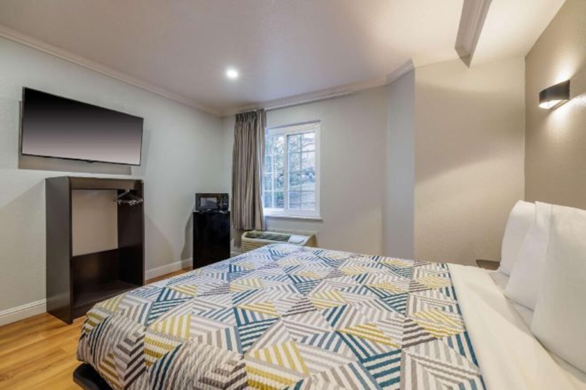 Habitación de hotel con cama, televisión montada en la pared, ventana con cortinas y una pequeña mesa con una silla.