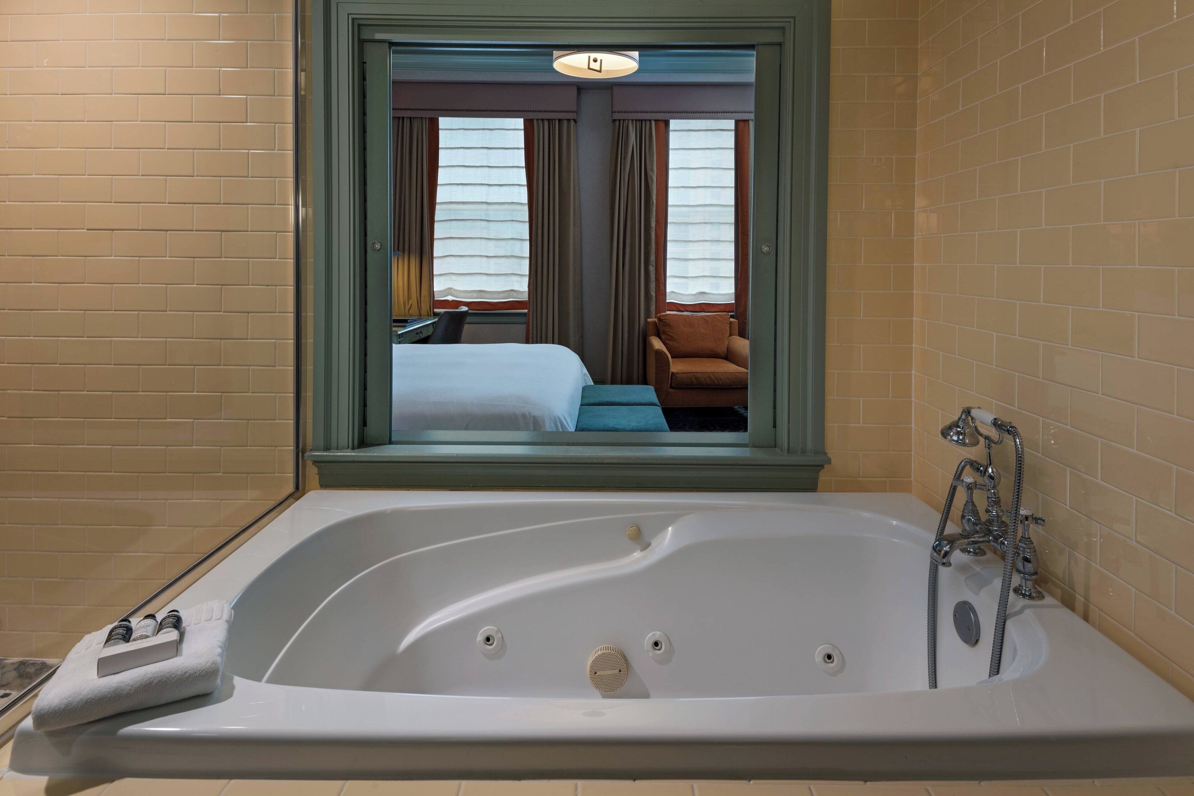 A hotel bathroom with a large bathtub, a window with a view of a bedroom, and a shower area.