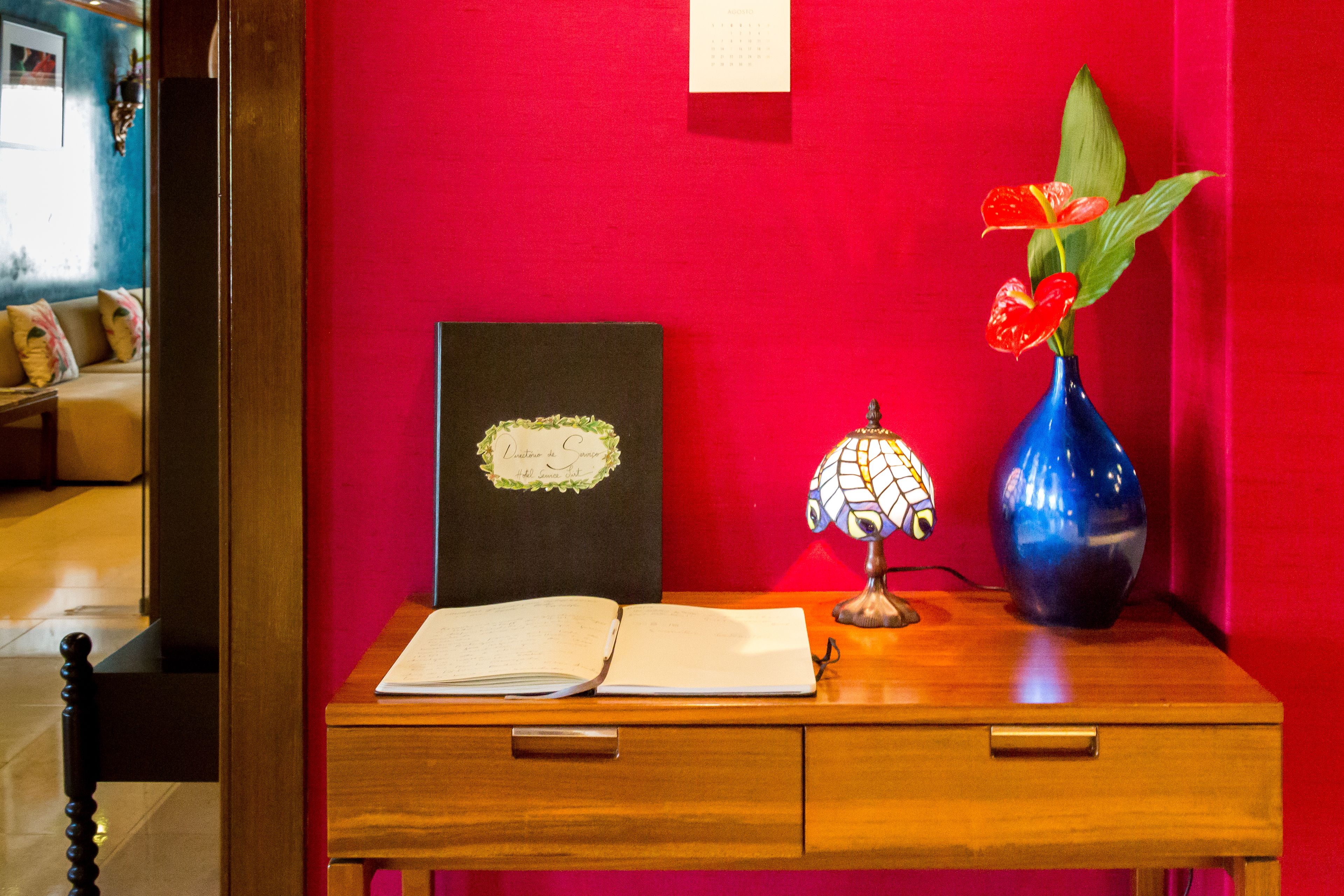 A wooden desk with a book, a framed picture, a lamp, and a vase with flowers against a red wall.