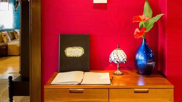 A wooden desk with a book, a framed picture, a lamp, and a vase with flowers against a red wall.