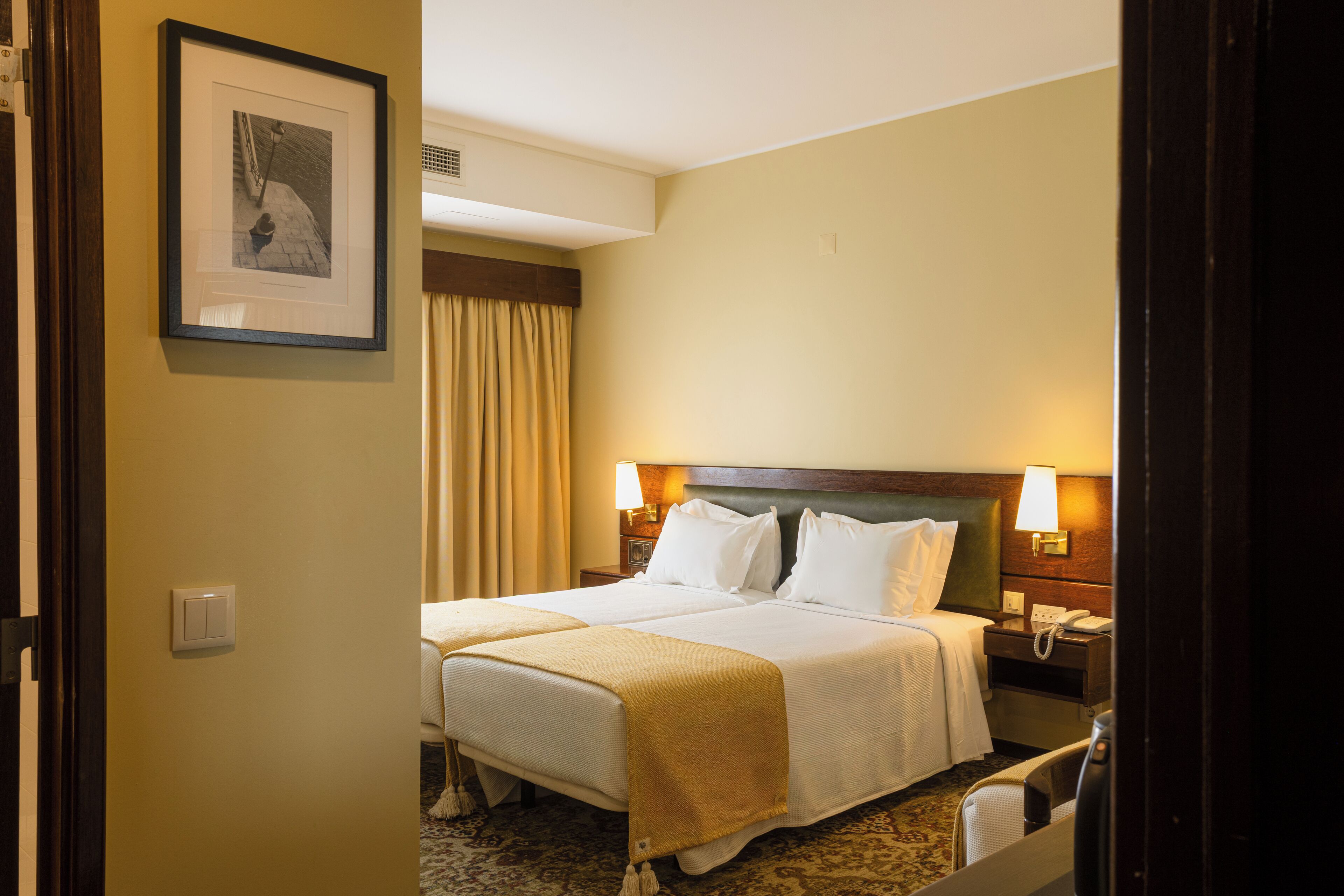 A hotel room with a double bed, bedside lamps, a nightstand, a telephone, and a framed picture on the wall.