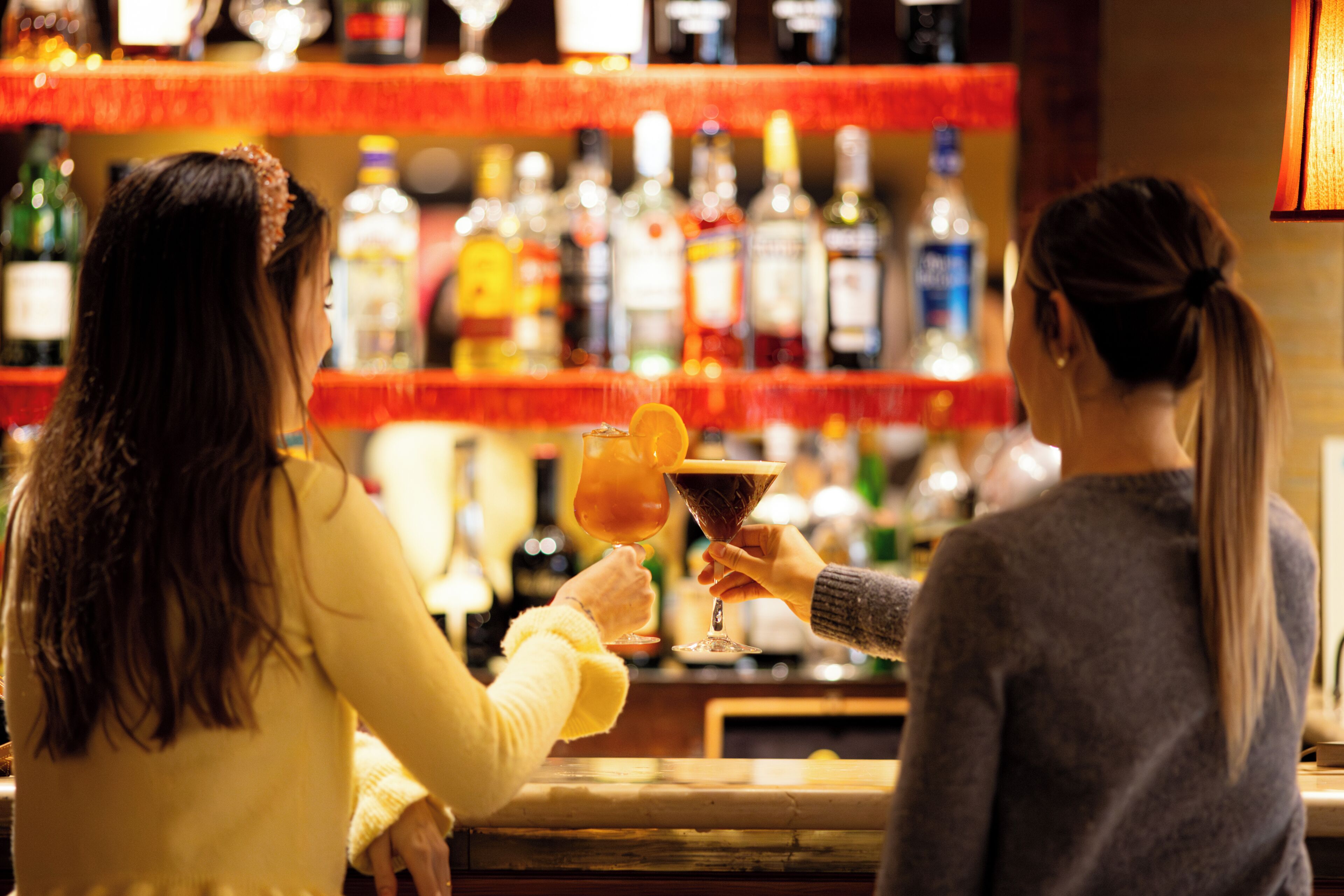 Dos personas brindando con bebidas en un bar con estantes llenos de botellas al fondo.