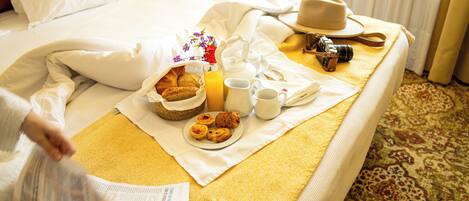 Una cama bien tendida con una bandeja de desayuno que incluye croissants, zumo de naranja y café, una cámara y un sombrero sobre la cama.