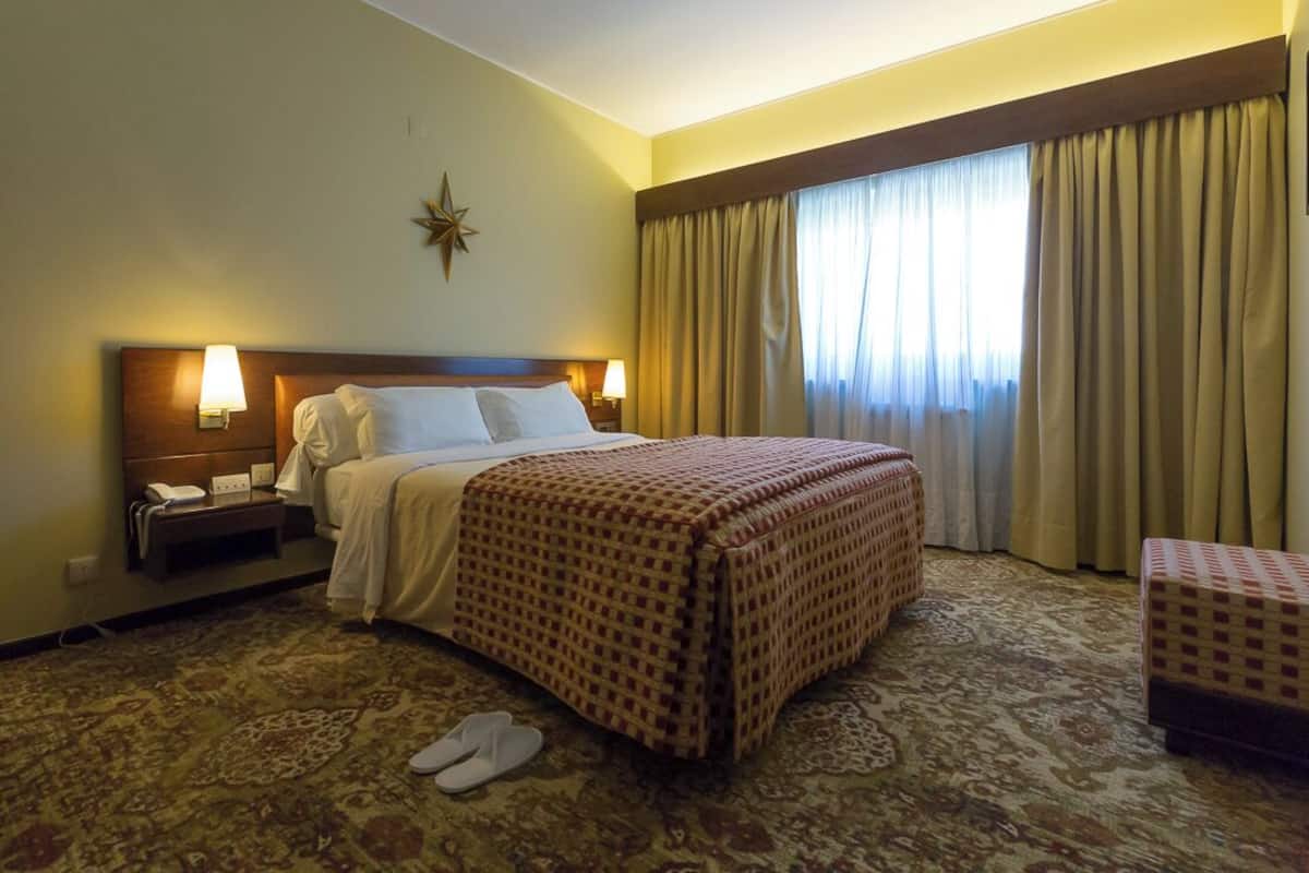 A hotel room with a bed, bedside lamps, a star decoration, a window with curtains, and a patterned carpet.