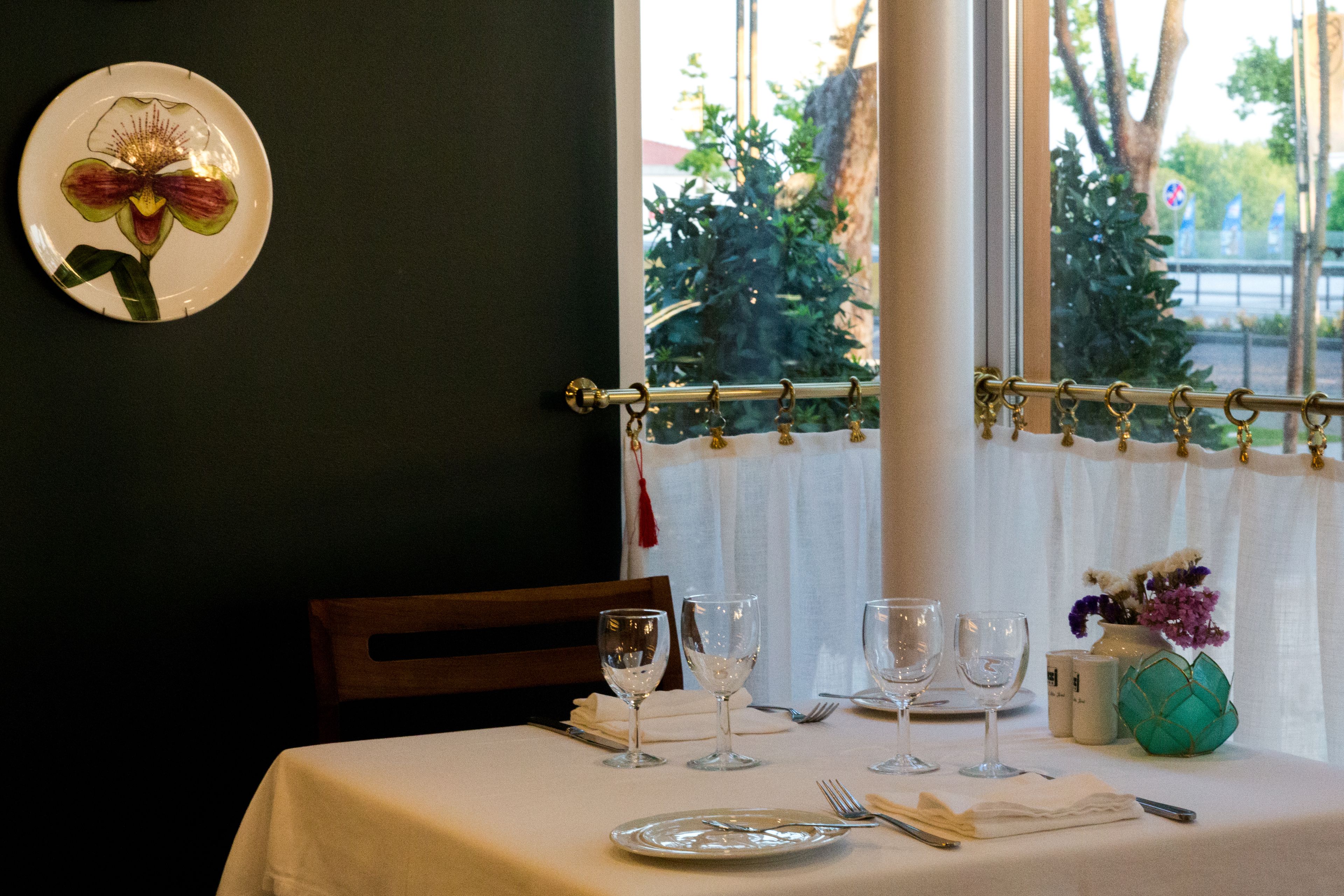 Een eettafel gedekt met borden, bestek en wijnglazen, een vaas met bloemen en een bloempot met een orchidee aan de muur.