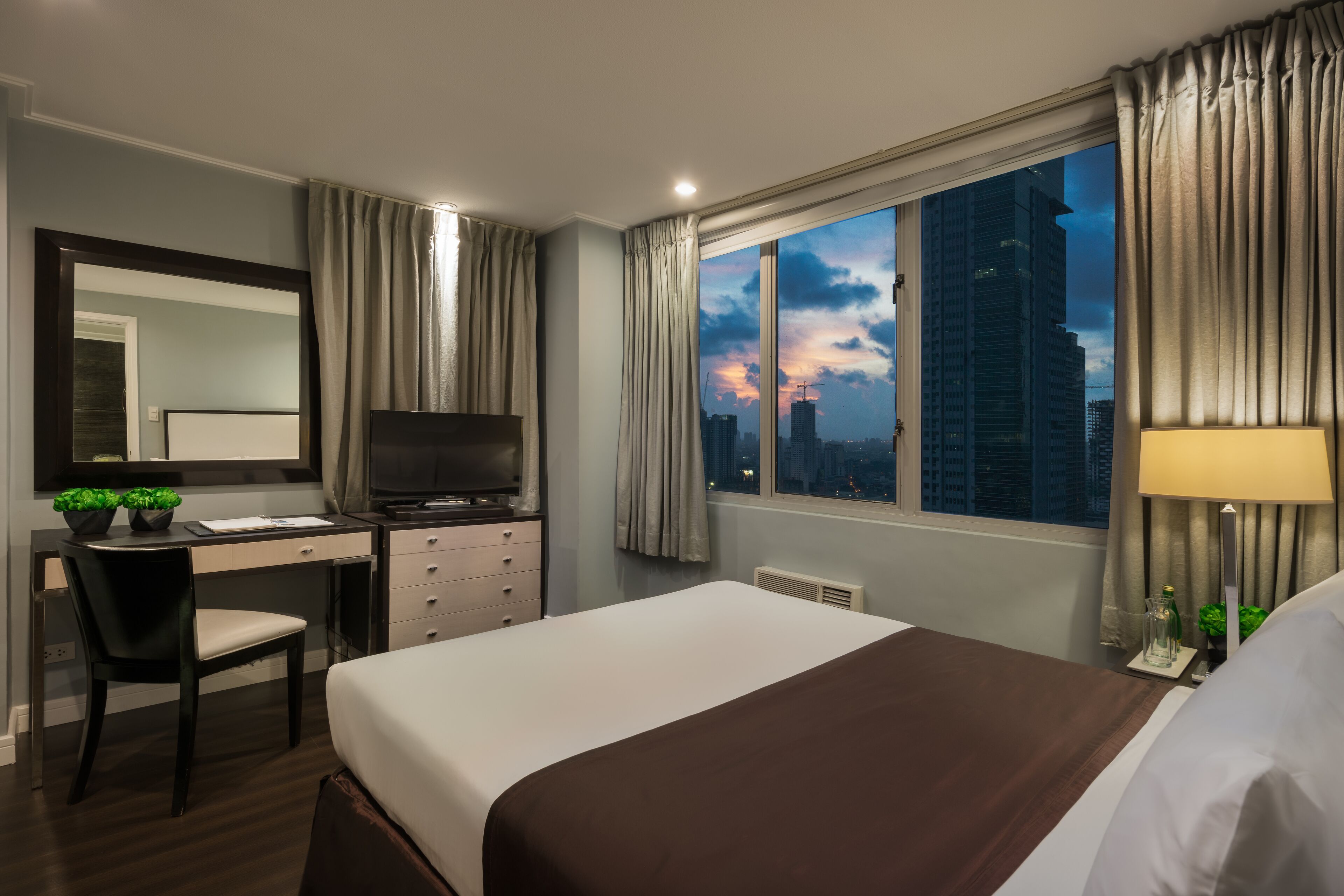 A hotel room with a bed, desk, chair, TV, and a large window with a city view.