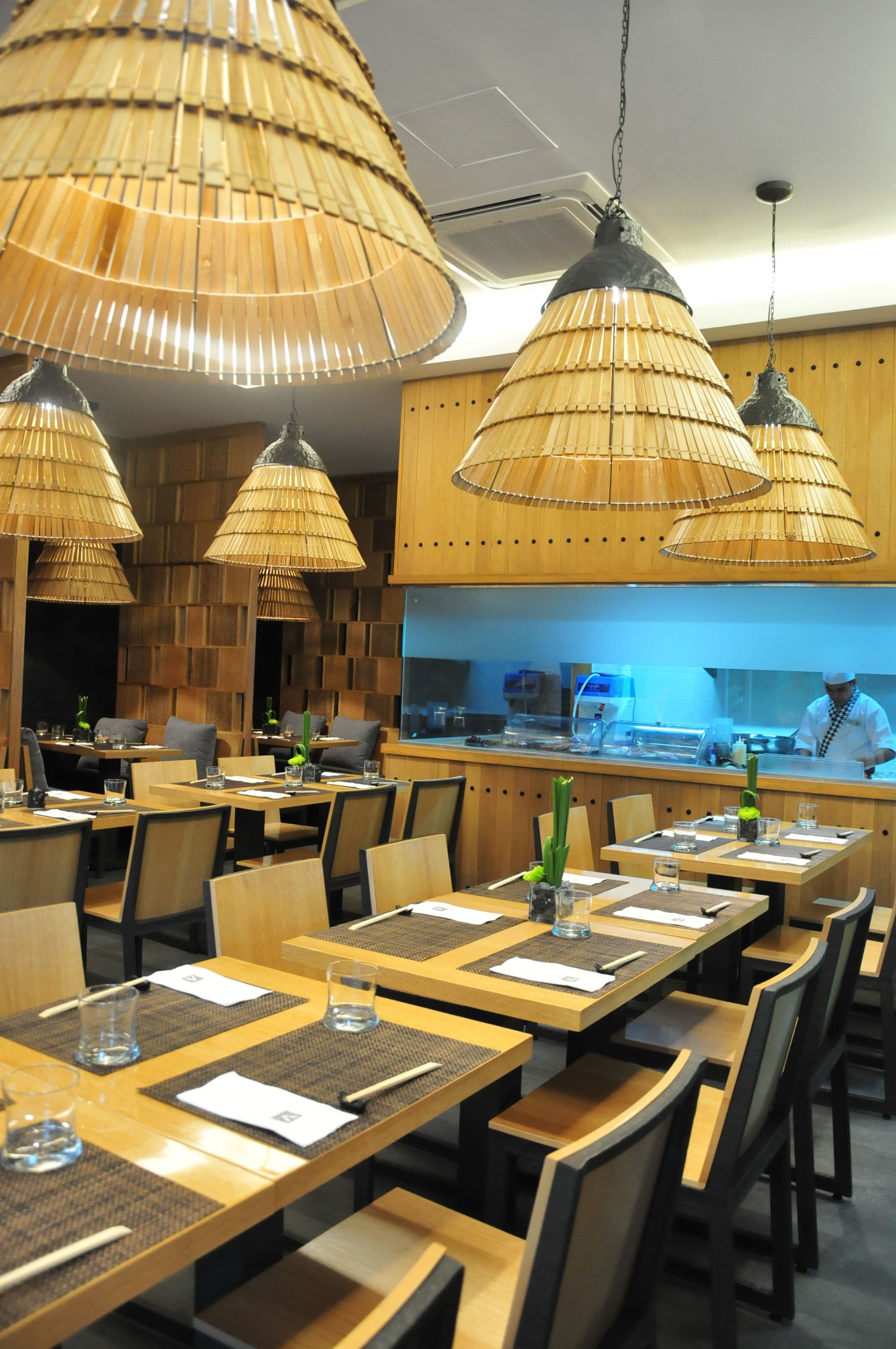 A restaurant with wooden tables and chairs, bamboo ceiling lamps, and a kitchen area in the background.