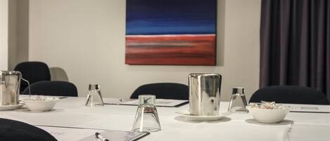 A conference room with a white table, chairs, a painting on the wall, and a window with curtains.