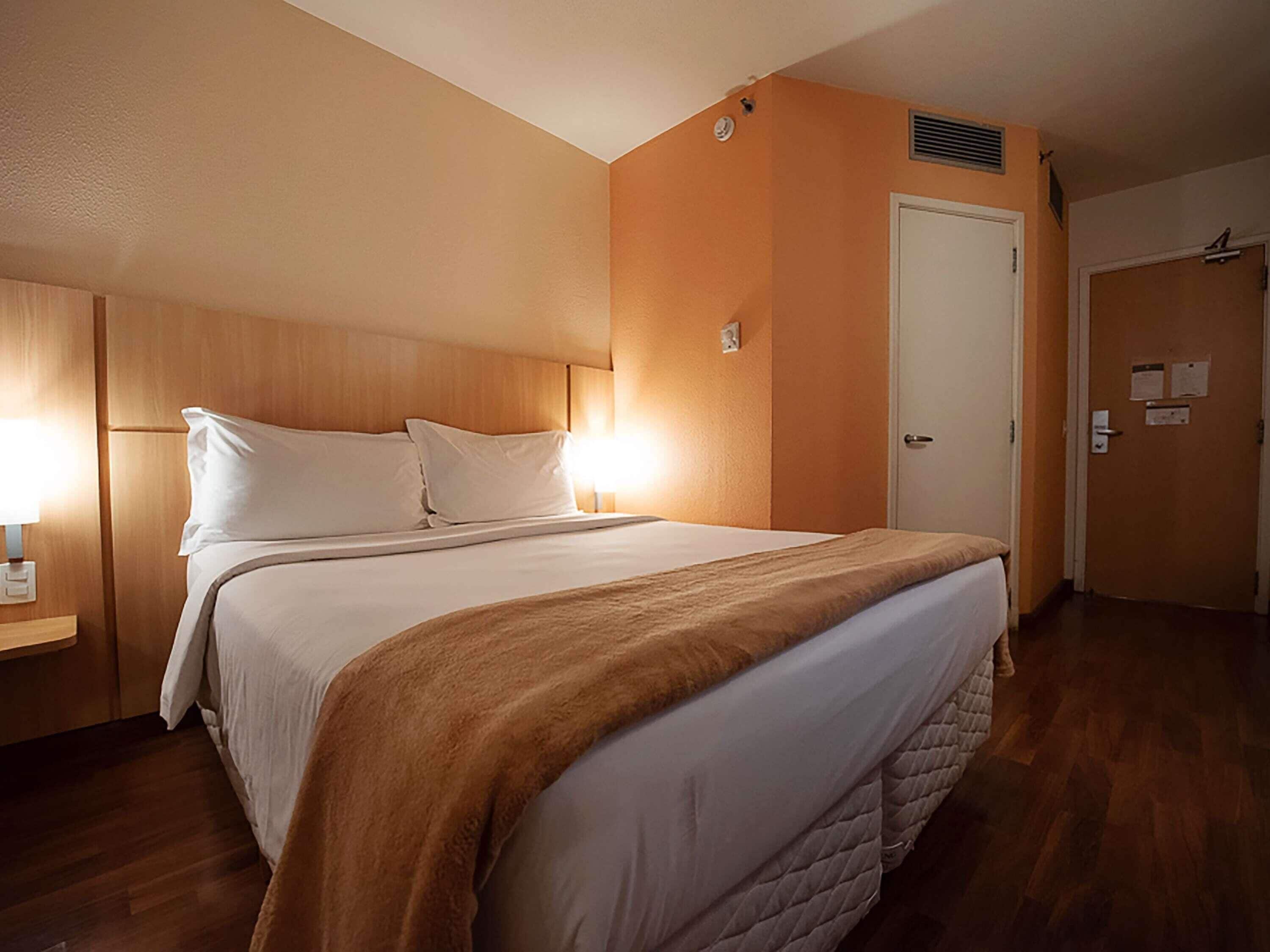 Quarto de hotel com cama, mesa de cabeceira, porta e luminária fixada na parede.