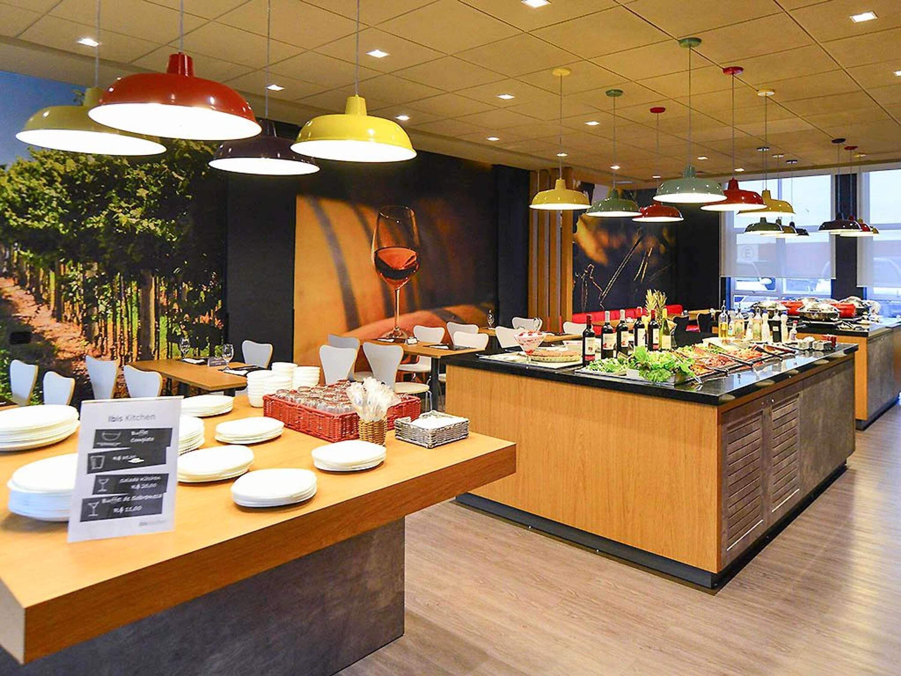 Un área de bufé con diversos alimentos, un cartel que indica 'Ibis Kitchen' y múltiples lámparas colgantes.