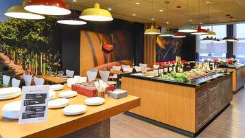 Un área de bufé con diversos alimentos, un cartel que indica 'Ibis Kitchen' y múltiples lámparas colgantes.