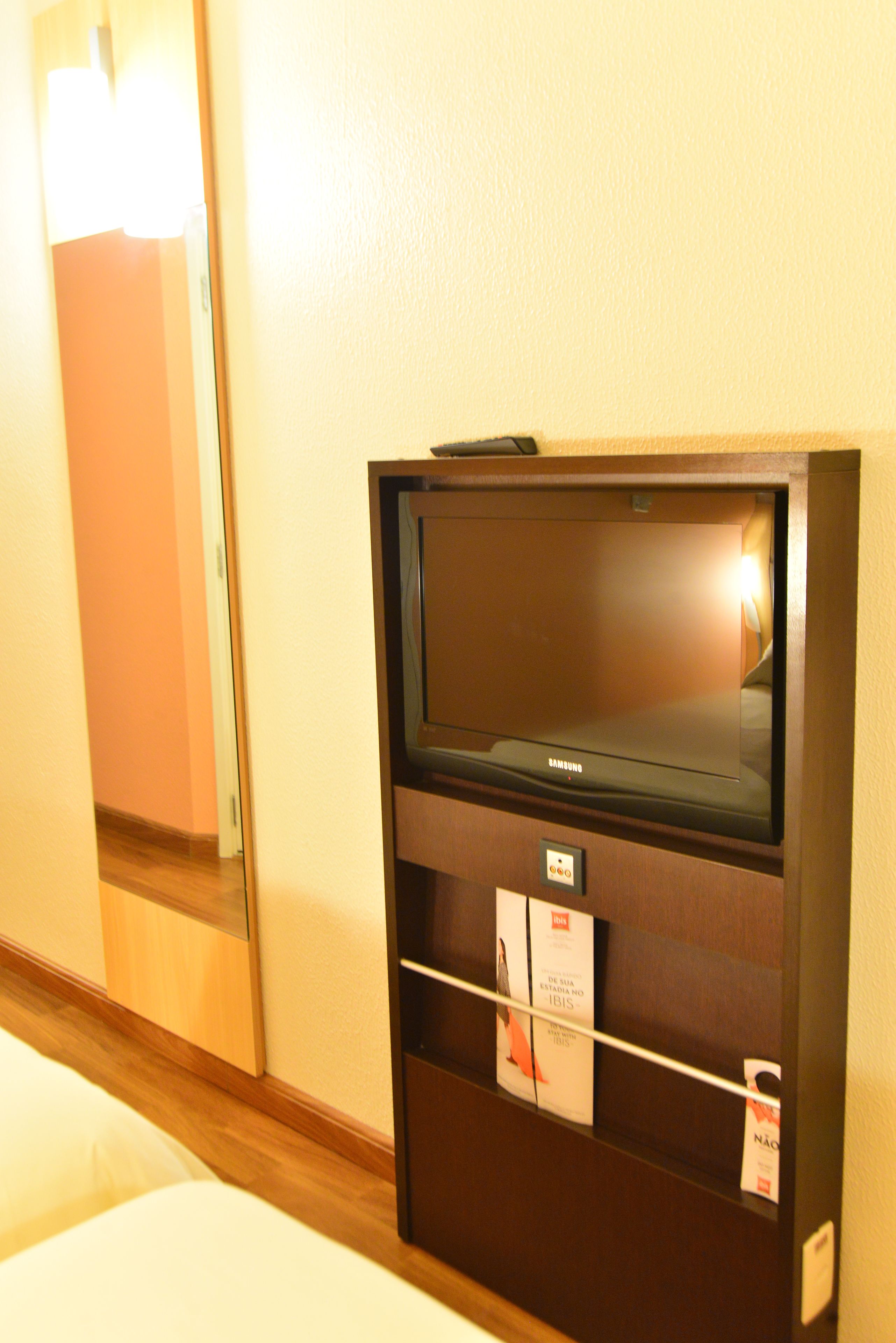 Quarto de hotel com uma cama, uma televisão presa à parede e uma pequena prateleira com objetos.