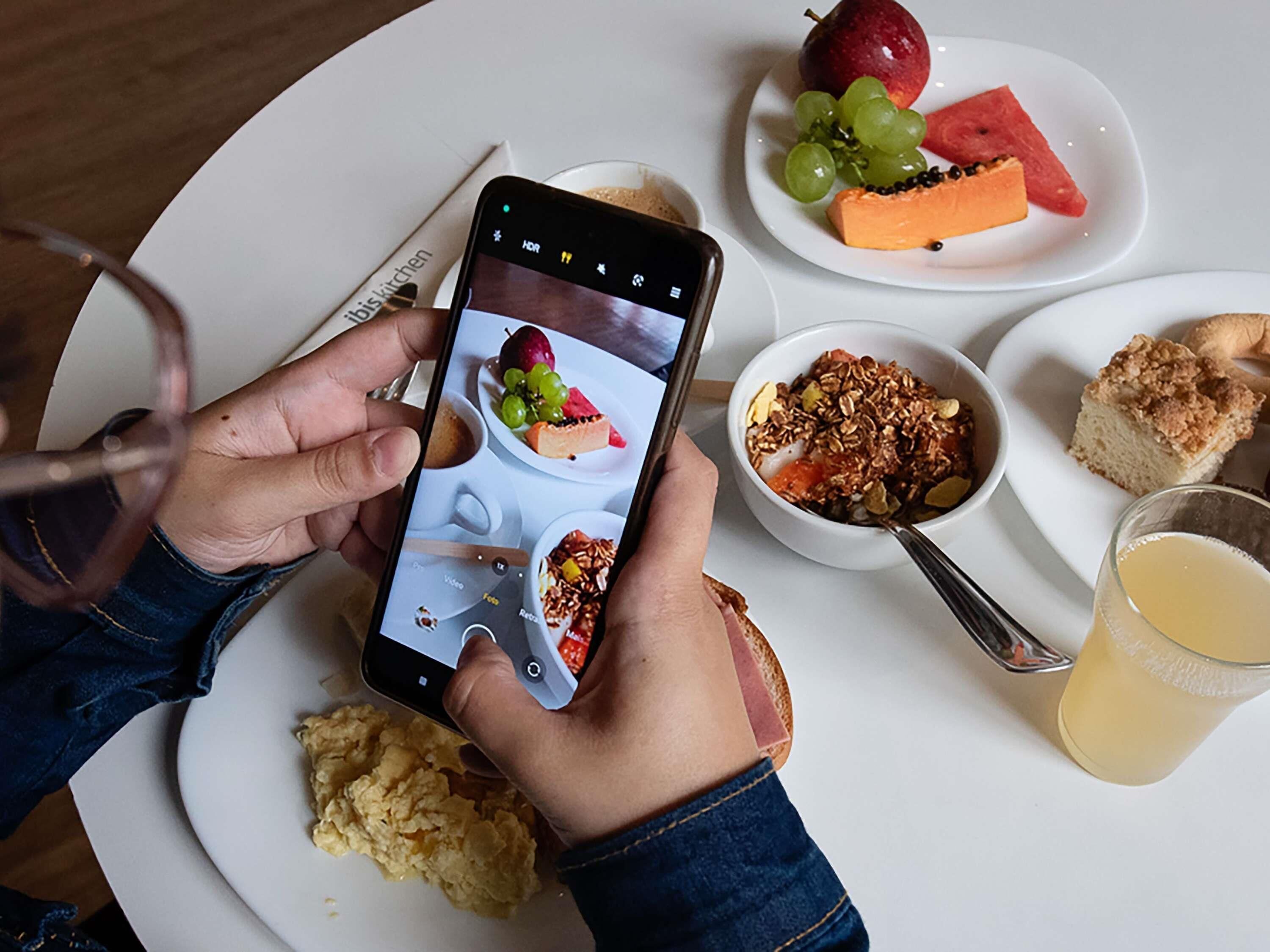 Eine Person hält ein Smartphone mit einer Essens-App in der Hand, auf dem Tisch ein Teller mit Rührei, eine Schüssel mit Granola, ein Glas Saft sowie ein Teller mit Obst und ein Stück Kuchen.