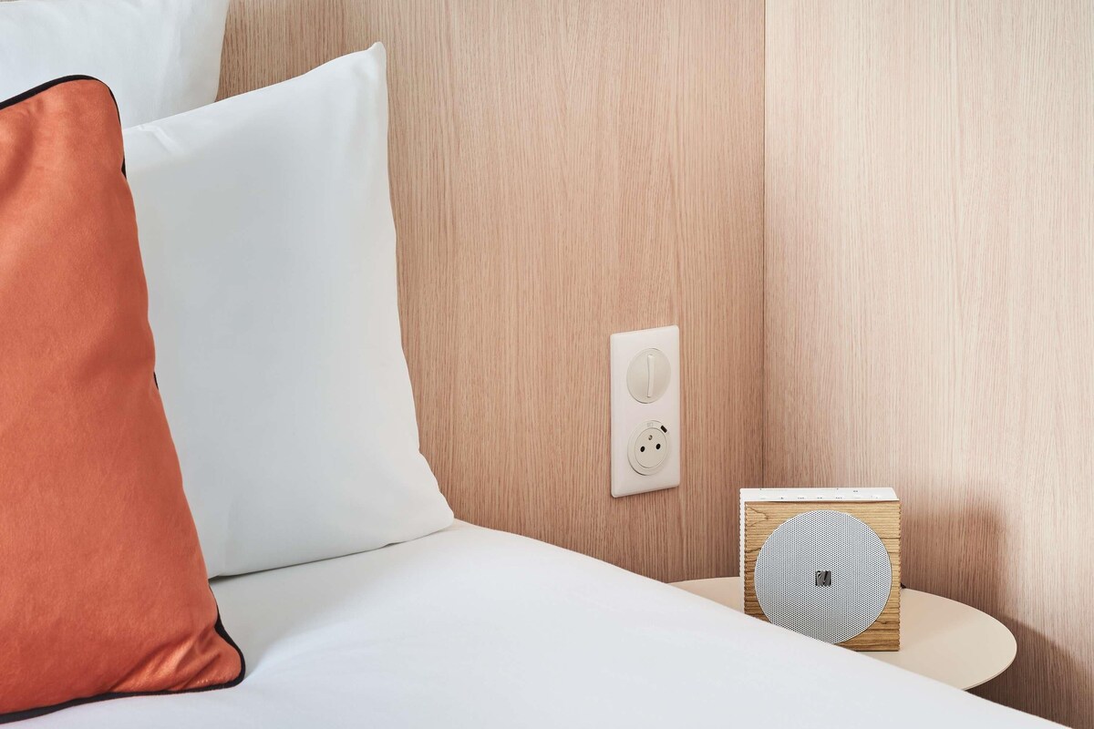 A neatly made bed with white and orange pillows, a power outlet, and a small wooden speaker on a bedside table.