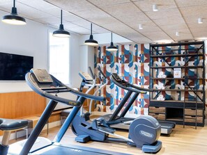 Een fitnessruimte met loopbanden, elliptische trainers en een muur met een kleurrijk abstract patroon.