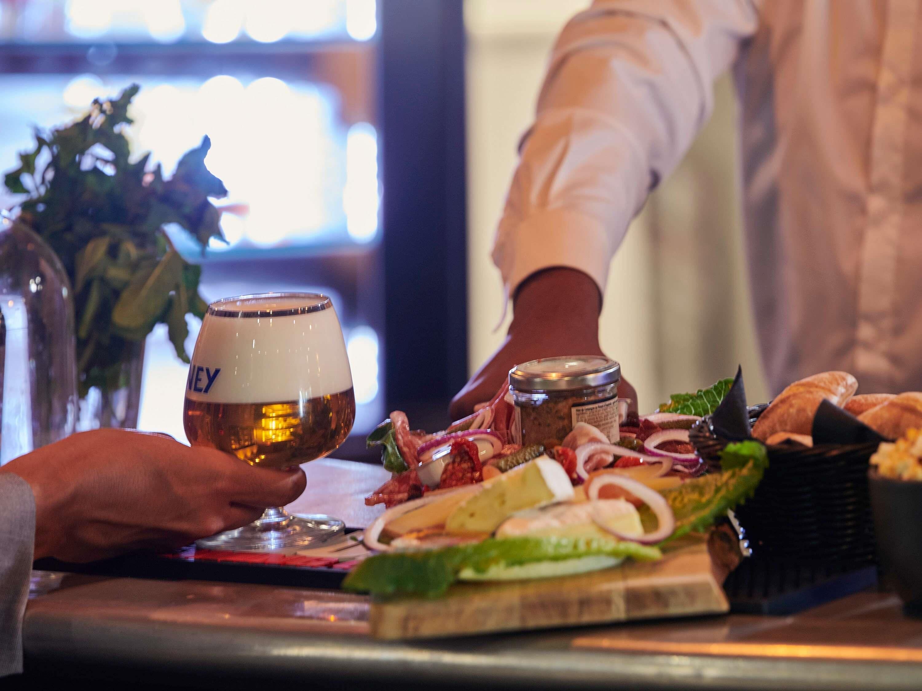 A person holding a glass of beer at a bar with a variety of food items and a bottle of beer on the counter.