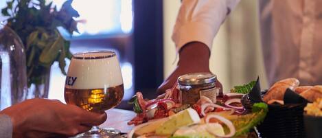 A person holding a glass of beer at a bar with a variety of food items and a bottle of beer on the counter.