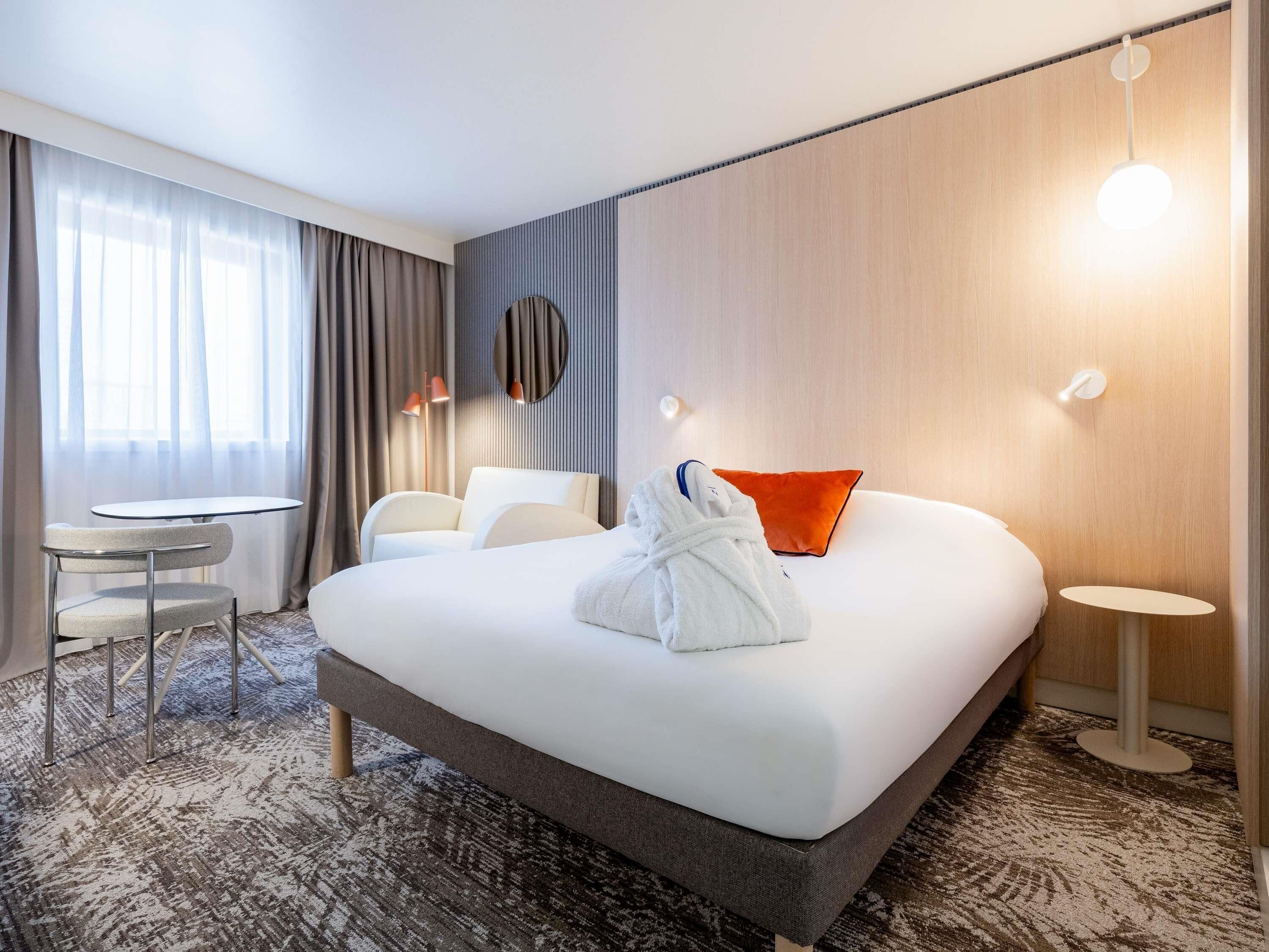 Habitación de hotel con una cama grande, una mesita redonda pequeña, una silla, una bata y una mesita de noche con lámpara.