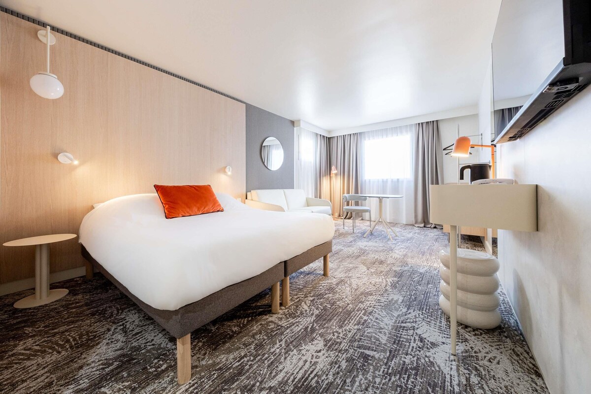 A modern hotel room with a large bed, a desk, a chair, and a small table.
