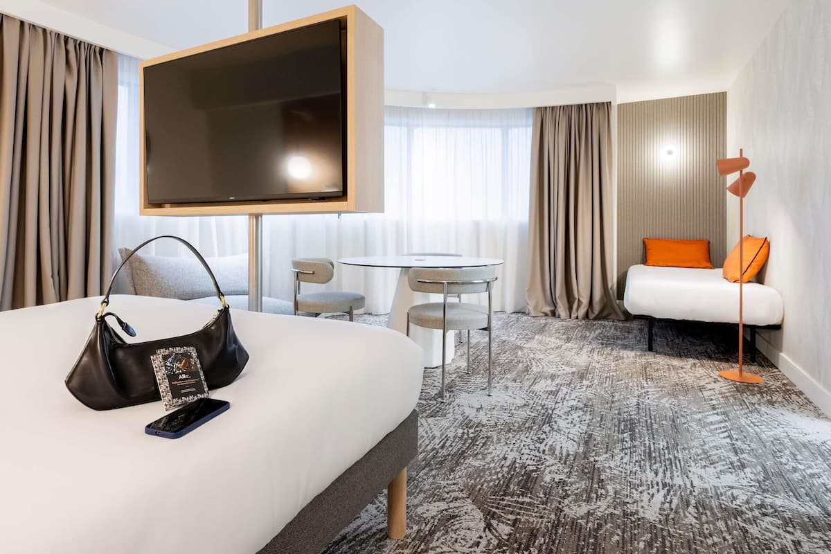 Una habitación de hotel con cama, televisor montado en la pared, escritorio con sillas, sofá y lámpara de pie.
