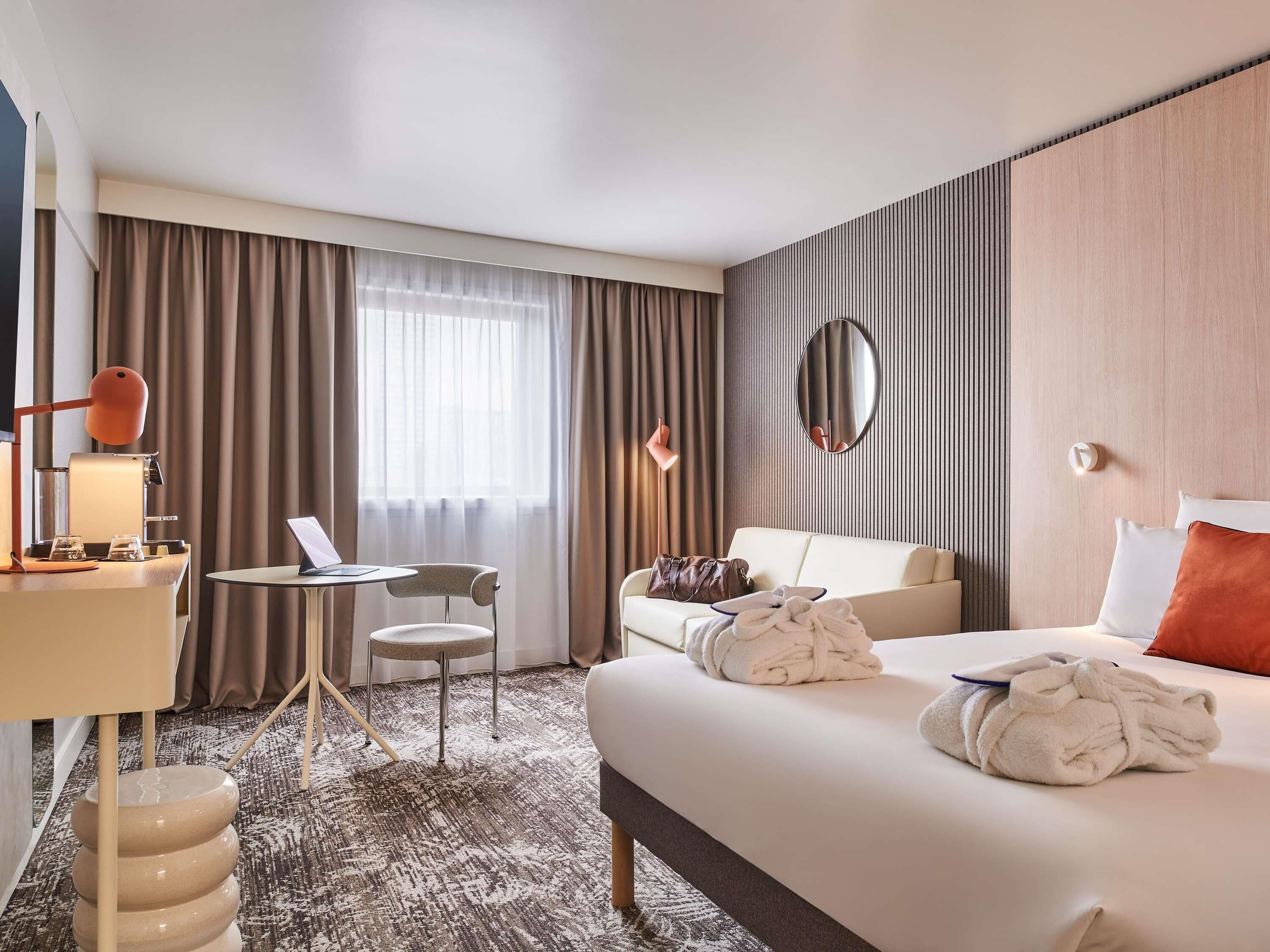 Une chambre d’hôtel avec un lit, un bureau, une chaise, un miroir rond et une fenêtre avec des rideaux.