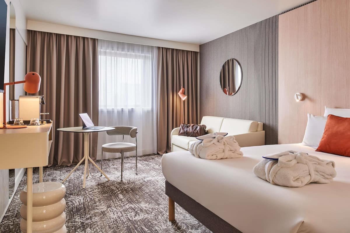 Une chambre d’hôtel avec un lit, un bureau, une chaise, un miroir rond et une fenêtre avec des rideaux.