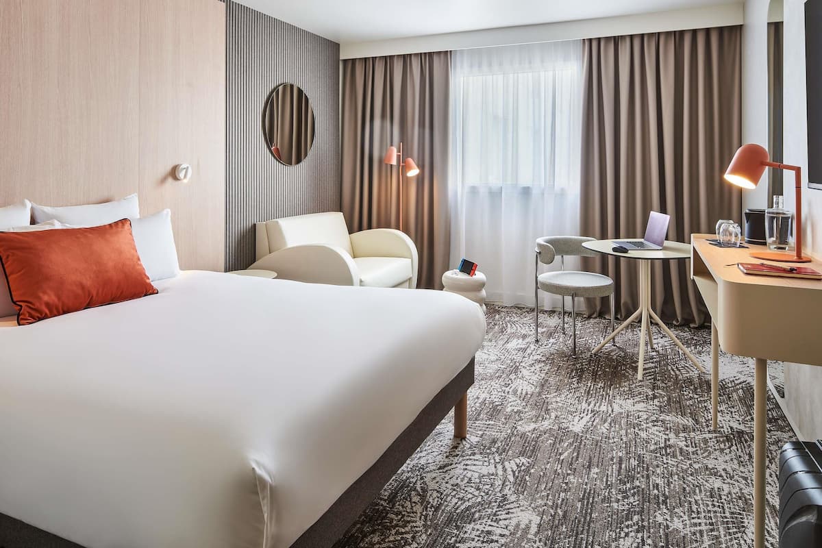 Habitación de hotel con una cama grande, un escritorio con una computadora portátil, una silla, un espejo redondo y vistas al exterior a través de la ventana.