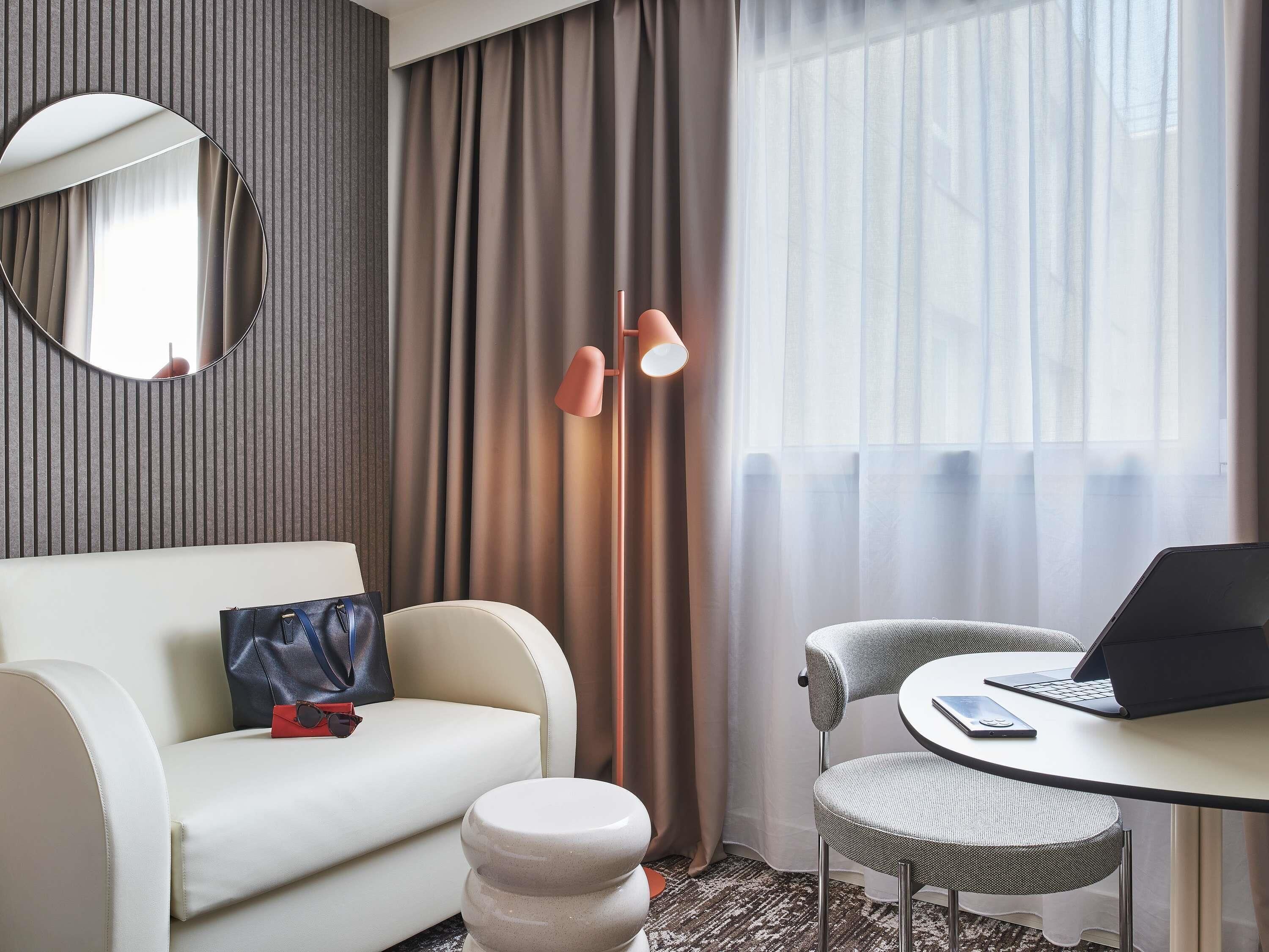 Een moderne hotelkamer met een sofa, een klein rond tafeltje met een laptop, een stoel, een lamp en een spiegel.